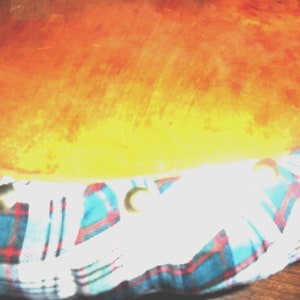 May include: A close-up of a wooden drum with a plaid fabric cover. The fabric is blue, white, and red with a tartan pattern. The drum is covered in a thin layer of dust.