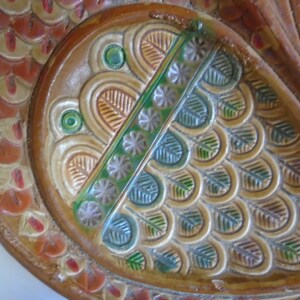 May include: A close-up of a decorative ceramic plate with a brown, orange, and green design. The plate features a stylized fish design with scales and a crown.