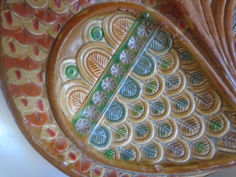 May include: A close-up of a decorative ceramic plate with a brown, orange, and green design. The plate features a stylized fish design with scales and a crown.