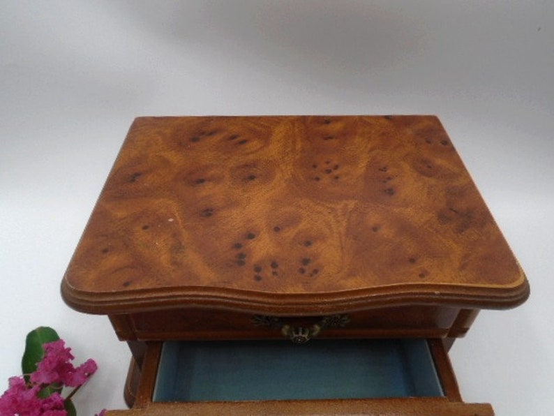 Peut inclure: Une bo&icirc;te &agrave; bijoux en bois avec un seul tiroir. La bo&icirc;te est de couleur brun clair avec un motif de grain de bois. Le tiroir est doubl&eacute; de bleu.