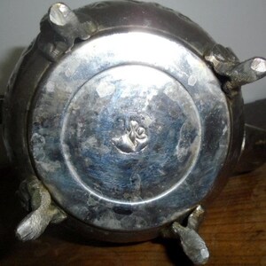 Puede incluir: Tetera de plata con detalles ornamentados y un dise&ntilde;o floral en la parte inferior. La tetera tiene cuatro patas y un pico.