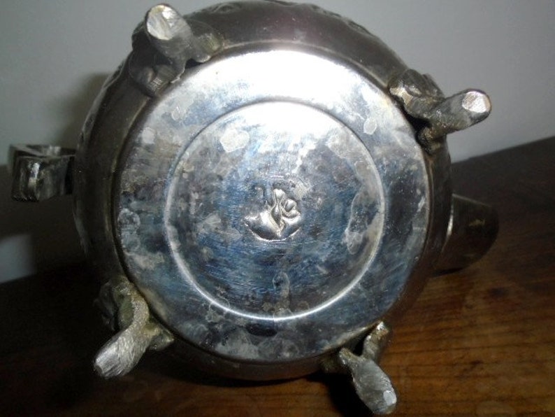 Puede incluir: Tetera de plata con detalles ornamentados y un dise&ntilde;o floral en la parte inferior. La tetera tiene cuatro patas y un pico.