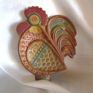 May include: A ceramic rooster wall decor with intricate details. The rooster is painted in shades of red, green, yellow, and brown. The rooster's tail feathers are spread out and have a leafy design.