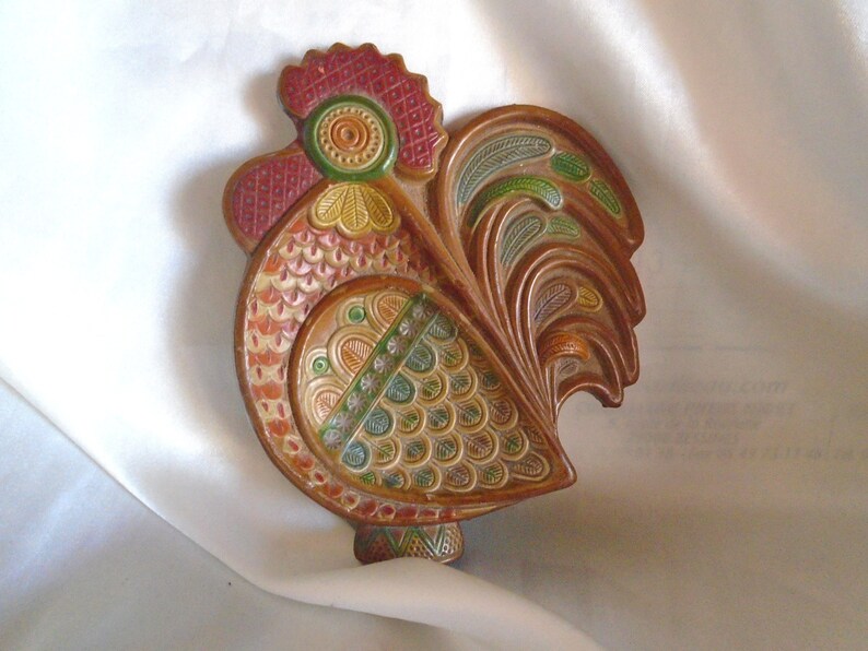 May include: A ceramic rooster wall decor with intricate details. The rooster is painted in shades of red, green, yellow, and brown. The rooster's tail feathers are spread out and have a leafy design.