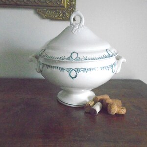 May include: A white porcelain tureen with a green and white floral design. The tureen has two handles and a lid. It is sitting on a wooden table with several wine corks.