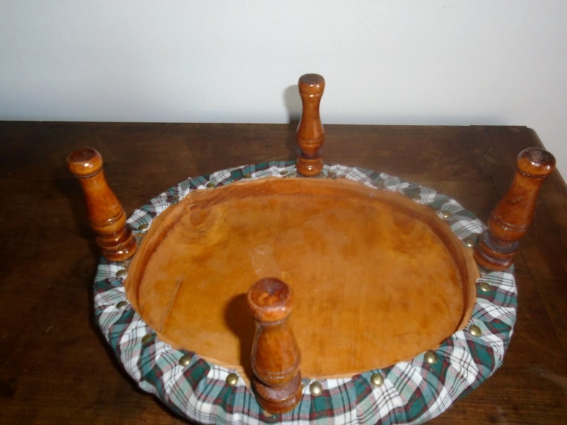 May include: The underside of a round, wooden stool with four legs. The stool is upholstered in a green and white plaid fabric. The fabric is secured with brass tacks.