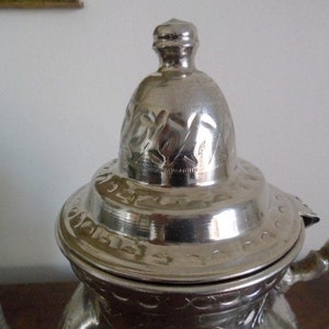 Puede incluir: Tetera de plata con una tapa adornada y un asa decorativa. La tapa tiene grabados intrincados y el cuerpo de la tetera est&aacute; decorado con un patr&oacute;n repetitivo.