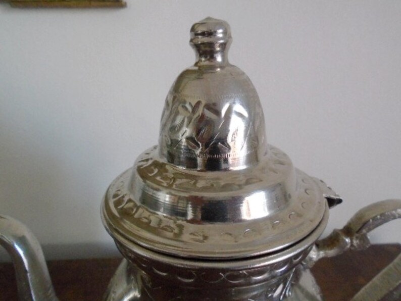 Puede incluir: Tetera de plata con una tapa adornada y un asa decorativa. La tapa tiene grabados intrincados y el cuerpo de la tetera est&aacute; decorado con un patr&oacute;n repetitivo.