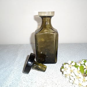 May include: A dark green glass bottle with a square body and a round stopper. The bottle is empty and the stopper is off.