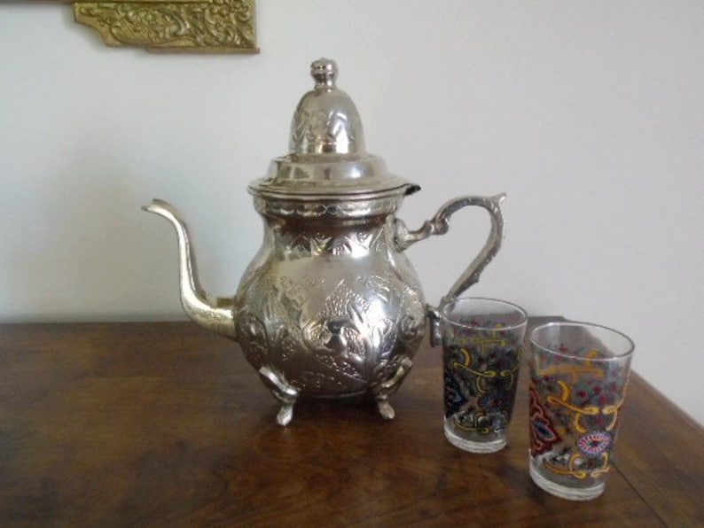 Puede incluir: Una tetera de plata con un dise&ntilde;o adornado y una tapa, junto con dos tazas de vidrio de colores. La tetera tiene un pico y una manija. Las tazas tienen un patr&oacute;n floral.