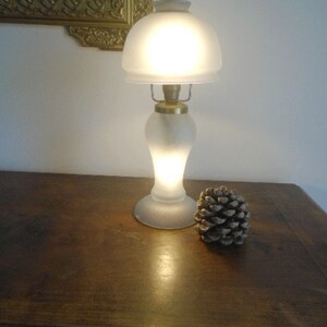 May include: A frosted glass table lamp with a gold base and a white shade. The lamp is turned on and illuminating the room. A pine cone is sitting on a wooden table next to the lamp.
