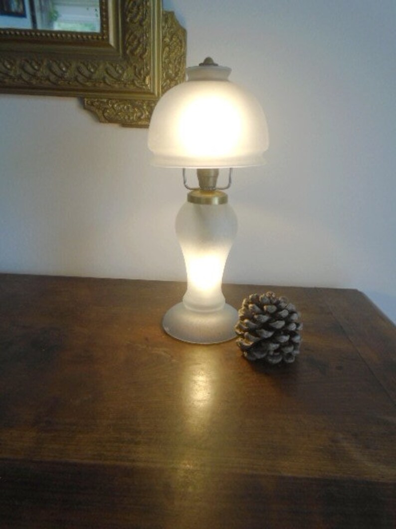 May include: A frosted glass table lamp with a gold base and a white shade. The lamp is turned on and illuminating the room. A pine cone is sitting on a wooden table next to the lamp.