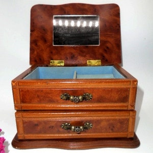 Peut inclure: Une bo&icirc;te &agrave; bijoux en bois avec deux tiroirs et un miroir &agrave; l'int&eacute;rieur. La bo&icirc;te est en bois brun clair avec un motif de grain de bois brun fonc&eacute;. Les tiroirs ont des poign&eacute;es en laiton.