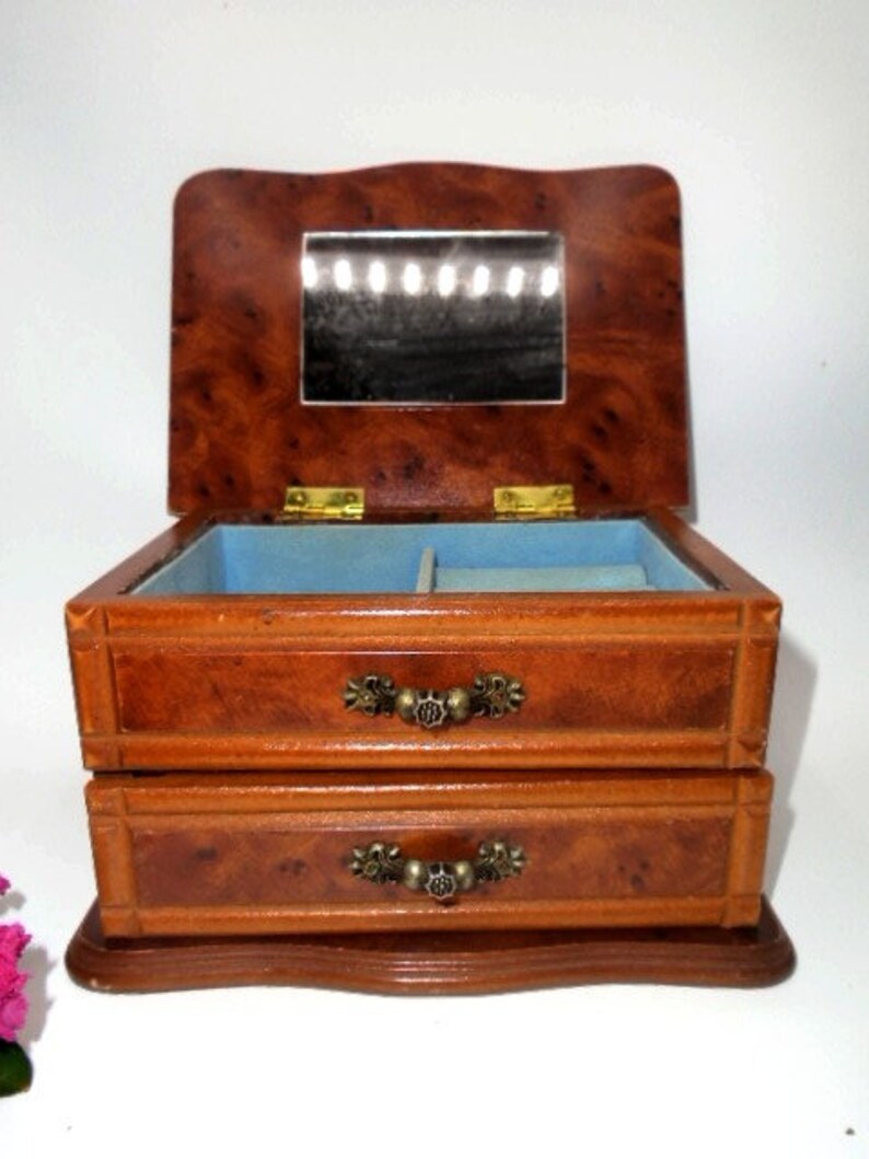 Peut inclure: Une bo&icirc;te &agrave; bijoux en bois avec deux tiroirs et un miroir &agrave; l'int&eacute;rieur. La bo&icirc;te est en bois brun clair avec un motif de grain de bois brun fonc&eacute;. Les tiroirs ont des poign&eacute;es en laiton.
