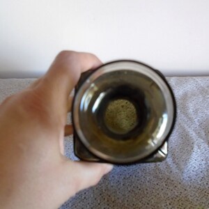 May include: A clear glass bottle with a dark green tint and a square shape. The bottle has a wide opening at the top.