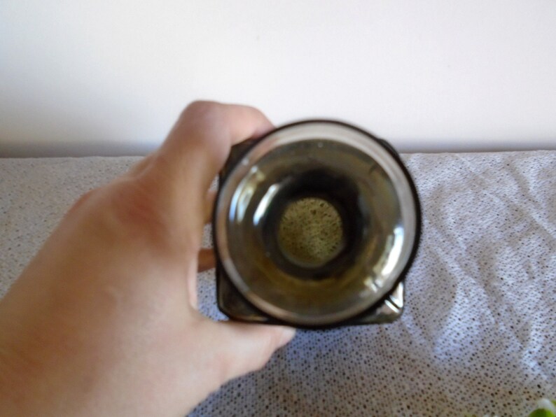 May include: A clear glass bottle with a dark green tint and a square shape. The bottle has a wide opening at the top.