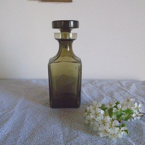 May include: A dark brown glass decanter with a square body and a round stopper. The decanter is sitting on a white tablecloth with a floral pattern.