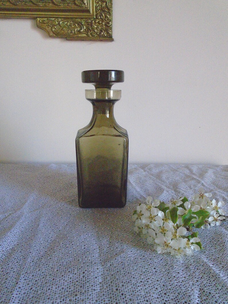 May include: A dark brown glass decanter with a square body and a round stopper. The decanter is sitting on a white tablecloth with a floral pattern.