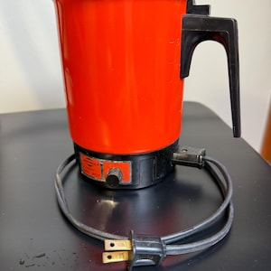 May include: A retro electric coffee pot in a vibrant red hue, featuring a black handle and lid. The base is black, with a dial, and a power cord with a two-pin plug. The cord is coiled around the base of the pot.