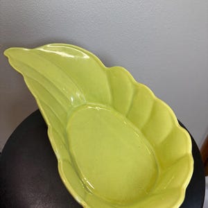 May include: A bright green, leaf-shaped ceramic dish. The dish has a scalloped edge and a smooth, glossy finish. The interior is oval-shaped and slightly recessed.