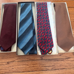 May include: Four neckties in open boxes. The ties are in shades of burgundy, blue, red, and brown. The burgundy tie is solid, the blue tie has vertical stripes, the red tie has a repeating oval pattern, and the brown tie is solid.