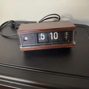 May include: A vintage Copal flip clock with a brown wood-grain finish. The clock displays the time as 5:10 PM in white numerals on black panels. The clock has a clear plastic cover and a black power cord.
