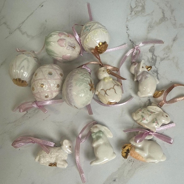 May include: A collection of ten ceramic ornaments featuring various shapes including eggs, a chick, a duck, and rabbits. Each ornament is finished in white with pastel accents, gold detailing, and includes a light pink ribbon for hanging purposes.