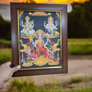 May include: A framed print of three Hindu deities, Lakshmi, Saraswati, and Ganesha, in a brown frame with a gold trim. The print is set against a black background and features intricate details and vibrant colors.