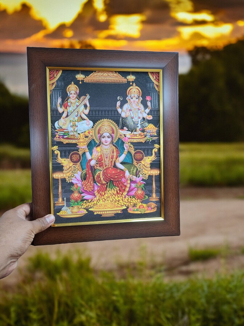 May include: A framed print of three Hindu deities, Lakshmi, Saraswati, and Ganesha, in a brown frame with a gold trim. The print is set against a black background and features intricate details and vibrant colors.