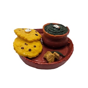 May include: A miniature clay model of a traditional Indian thali, featuring a small clay pot filled with green food, two round yellow flatbreads, and a small piece of ginger on a red clay plate.