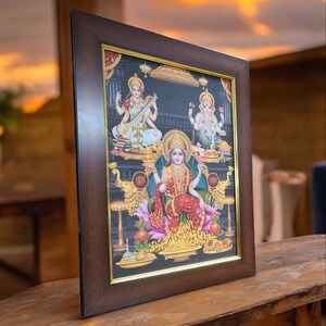 May include: A framed print of three Hindu deities. The central figure is a woman with a golden crown and a red and gold sari. She is surrounded by flowers and gold coins. The figures on either side are a woman playing a stringed instrument and a man with an elephant head. The frame is brown wood with a gold trim.