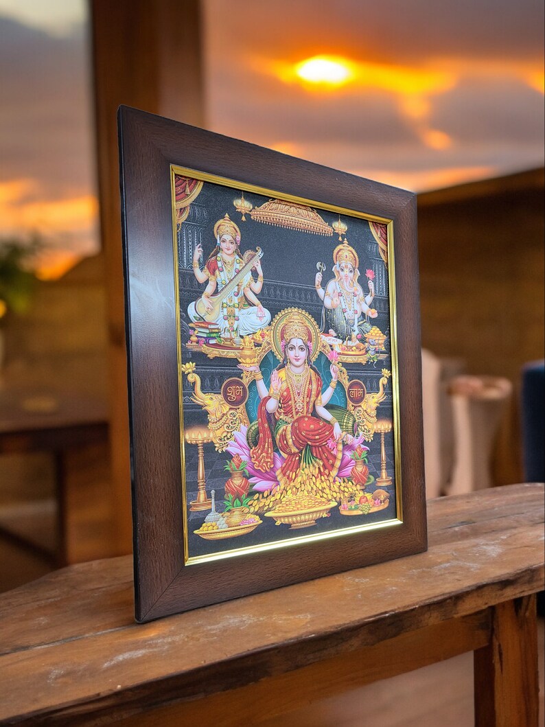 May include: A framed print of three Hindu deities. The central figure is a woman with a golden crown and a red and gold sari. She is surrounded by flowers and gold coins. The figures on either side are a woman playing a stringed instrument and a man with an elephant head. The frame is brown wood with a gold trim.