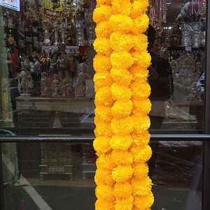 May include: A long garland of bright yellow marigold flowers, strung together with green string. The garland is hanging in front of a window with a reflection of a store interior.