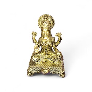 May include: A gold-colored statue of a seated deity, adorned with intricate details. The figure wears a crown and holds lotus flowers in two hands. The statue is on a decorative base, and the overall design is ornate.