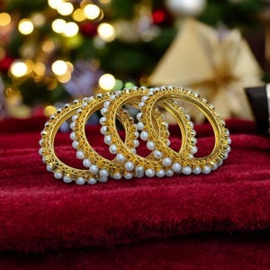 May include: Four gold bangles with white pearl accents. The bangles are stacked on top of each other on a red velvet surface.