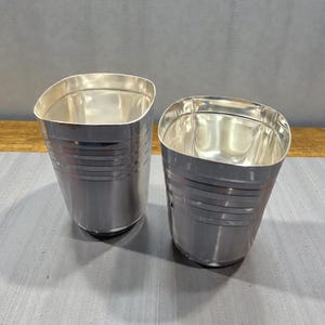 Puede incluir: Dos vasos de metal plateado con forma ligeramente curvada y rectangular. Cada vaso tiene líneas horizontales alrededor del cuerpo. El interior de los vasos refleja la luz. Los vasos están colocados sobre una superficie gris.