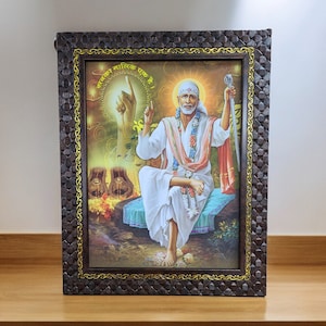 Puede incluir: Una imagen enmarcada de una figura religiosa, Sai Baba, con barba blanca y ropa blanca, sentado en un cojín azul. La imagen está rodeada por un marco de madera marrón con un borde dorado. El texto "सच्चर साधिक एक" es visible en la imagen.