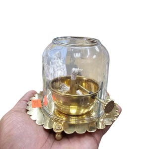 May include: A vintage brass oil lamp with a clear glass chimney. The lamp features a round base with a scalloped edge and a small handle. The wick is visible inside the brass bowl. A price tag is visible on the base.