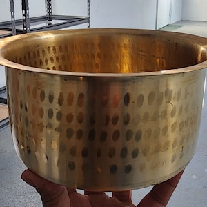 Könnte beinhalten: Eine große, runde, gehämmerte Schüssel aus Messing mit einem breiten Rand. Die Schüssel hat eine strukturierte Oberfläche mit einem Muster aus kleinen, runden Vertiefungen.