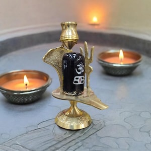 Op de afbeelding: Een religieus messing beeld met een zwarte cilindrische steen, versierd met witte symbolen. Het beeld staat op een decoratieve basis, met een cobra-achtig ontwerp. Twee brandende kaarsen in kleine kommen staan op de voorgrond.