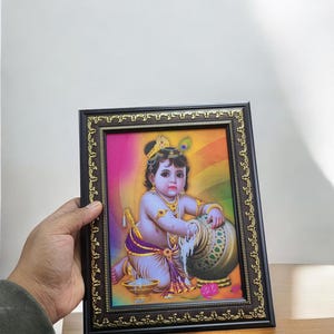 May include: A framed picture of a young boy, possibly Krishna, pouring milk from a pot. The picture is set against a colorful background with a rainbow and a golden halo around the boy's head. The frame is brown with a gold trim.