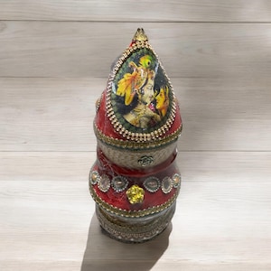 May include: A multi-tiered decorative box with a red, gold, and green design. The top tier features a printed image of a couple in traditional Indian clothing.