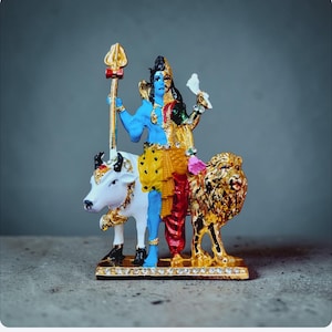 May include: A colorful figurine depicting a deity, half blue and half gold, holding a trident and a white bird. The figure stands on a gold base with a white bull and a golden lion. The figurine is a religious item.