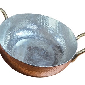 Puede incluir: Un bol de servir de aluminio martillado con dos asas de latón. El bol tiene un acabado plateado y una forma redondeada.
