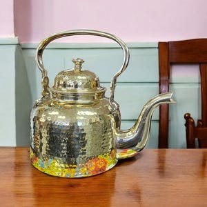 May include: A hammered brass teapot with a lid and a curved spout. The teapot is sitting on a wooden table.
