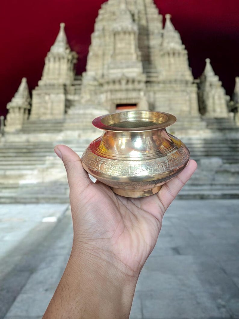 May include: A gold-colored metal pot with intricate designs sits in a hand, with a large, ornate, white temple in the background.