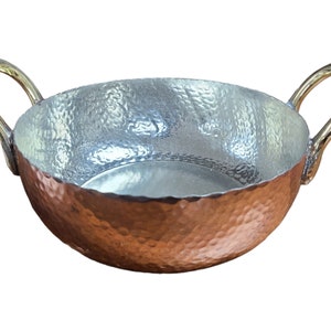 May include: A hammered copper serving bowl with two brass handles. The bowl has a rounded shape and a textured surface.