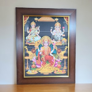May include: A framed print of a Hindu deity, Lakshmi, seated on a throne with two other deities, Saraswati and Ganesha, on either side. The print is set against a black background and features intricate details and vibrant colors. The text "शुभ" and "लाभ" is written in Hindi at the bottom of the print.