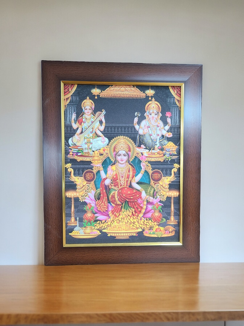 May include: A framed print of a Hindu deity, Lakshmi, seated on a throne with two other deities, Saraswati and Ganesha, on either side. The print is set against a black background and features intricate details and vibrant colors. The text "शुभ" and "लाभ" is written in Hindi at the bottom of the print.