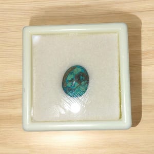 Pode incluir: Uma gema turquesa em cabochão em uma caixa de exibição forrada de espuma branca.